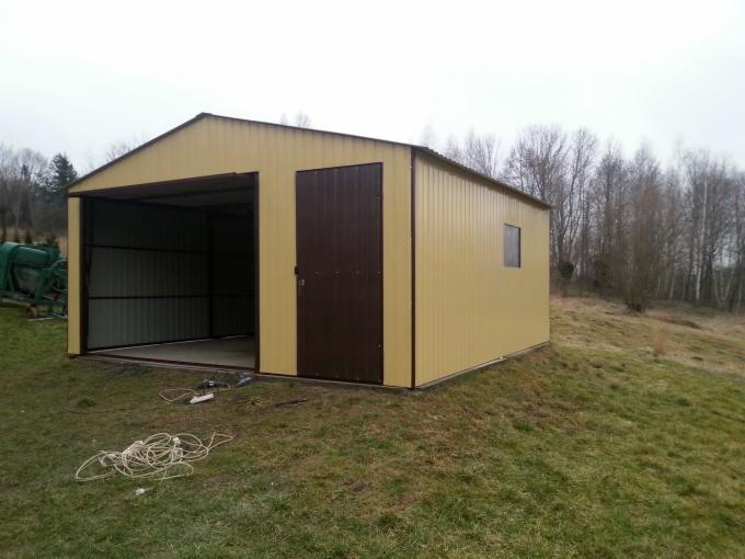 Żółty garaż blaszany z brązowymi drzwiami na zewnątrz, z widocznymi przewodami na trawie i zieloną maszyną w tle.