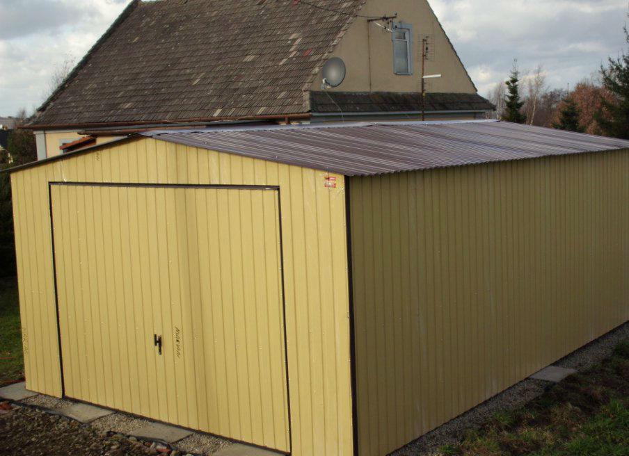 Żółty garaż blaszany z brązowym dachem na tle domu z brązowym dachem i satelitą. Widok z przodu, lekki kąt.
