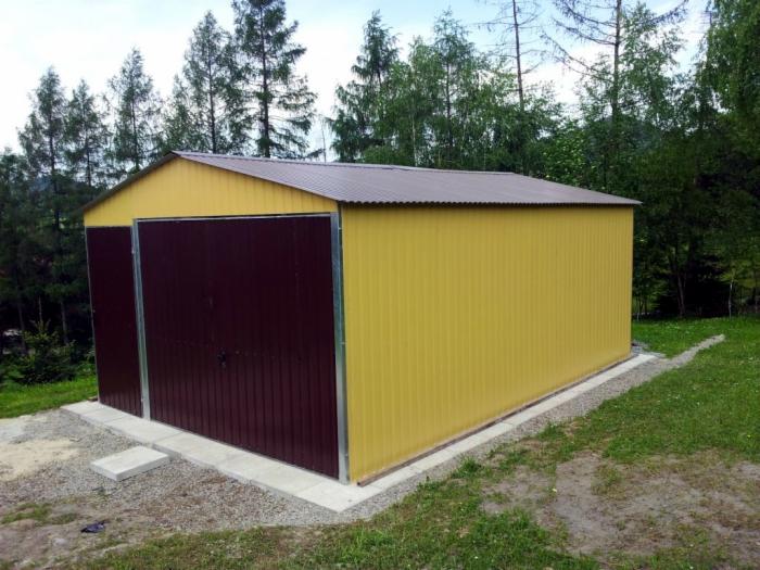 Żółty garaż blaszany z brązowymi drzwiami uchylnymi, widoczny na tle zielonego terenu z drzewami. Betonowa podstawa garażu. Ujęcie z lekkiej perspektywy.