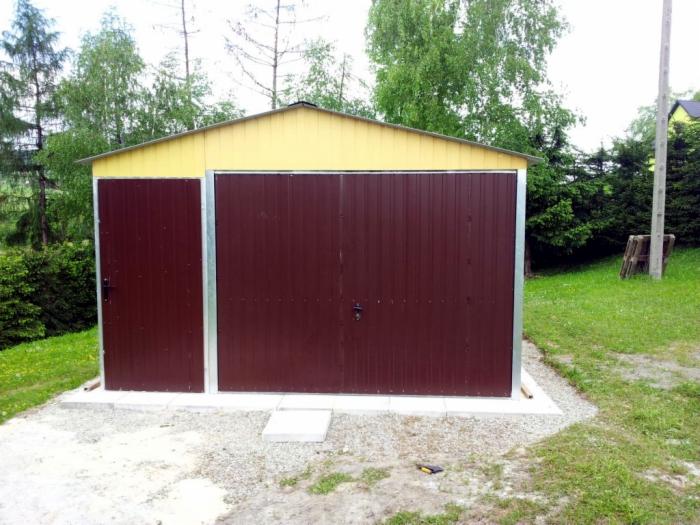 Gotowy garaż blaszany z brązowymi drzwiami i żółtym dachem, widoczny na tle zieleni. Mała furtka z boku. Garaż stoi na betonowej podstawie.