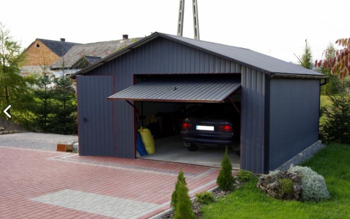 Grafitowy garaż blaszany z uchylnymi drzwiami, wewnątrz zaparkowany samochód. Kostka brukowa przed garażem, w tle budynek mieszkalny i zieleń.