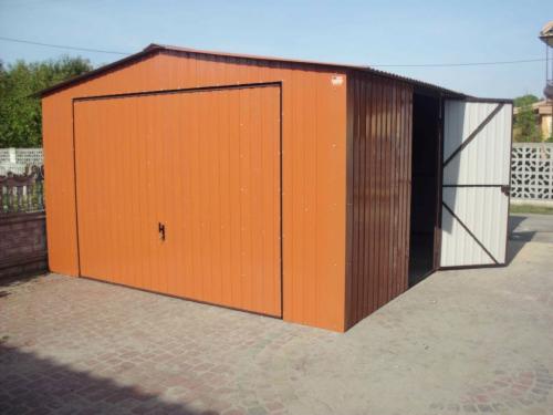 Pomarańczowo-brązowy garaż blaszany z otwartymi, dwuskrzydłowymi drzwiami bocznymi na brukowanym podłożu. Widok z boku, w tle zieleń.