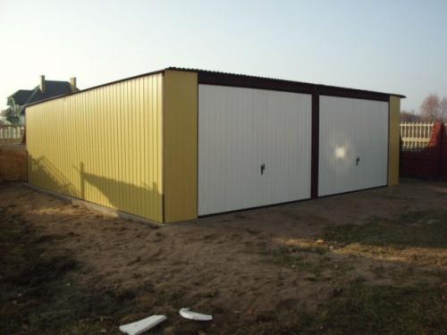 Żółto-biały garaż blaszany z dwiema bramami uchylnymi, widoczny z boku na tle ogrodu i domu. Garaż z blachy trapezowej, z brązowymi obramowaniami bram.