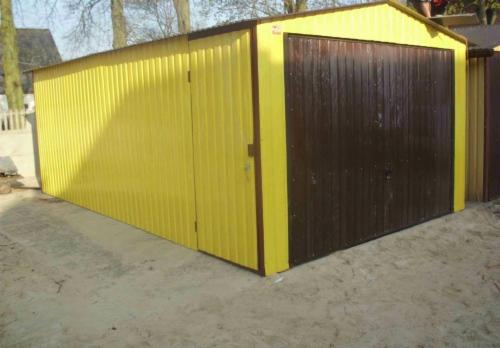 Żółty garaż blaszany z brązowymi drzwiami, widok z boku, na piaszczystym podłożu, w tle drzewa i fragment budynku. Widoczna boczna furtka.