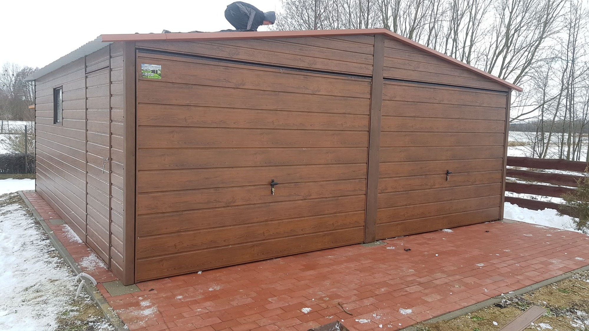 Brązowy garaż dwustanowiskowy z paneli imitujących drewno, z pracownikiem na dachu w trakcie montażu, na czerwonej kostce brukowej, zimowy krajobraz w tle.