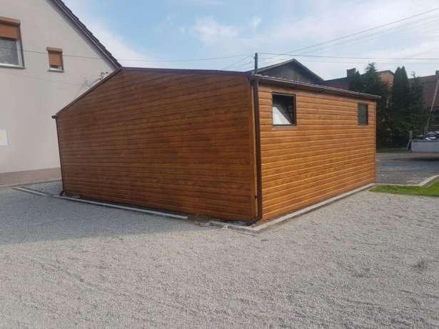 Gotowy, drewnopodobny garaż z dwoma małymi oknami na żwirowym podłożu, stojący obok domu.