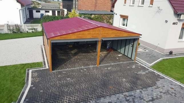 Dwustanowiskowy garaż z drewnianą konstrukcją, czerwoną blachodachówką i szarymi ścianami, wyłożony kostką brukową na posesji z trawnikiem i budynkiem mieszkalnym w tle.