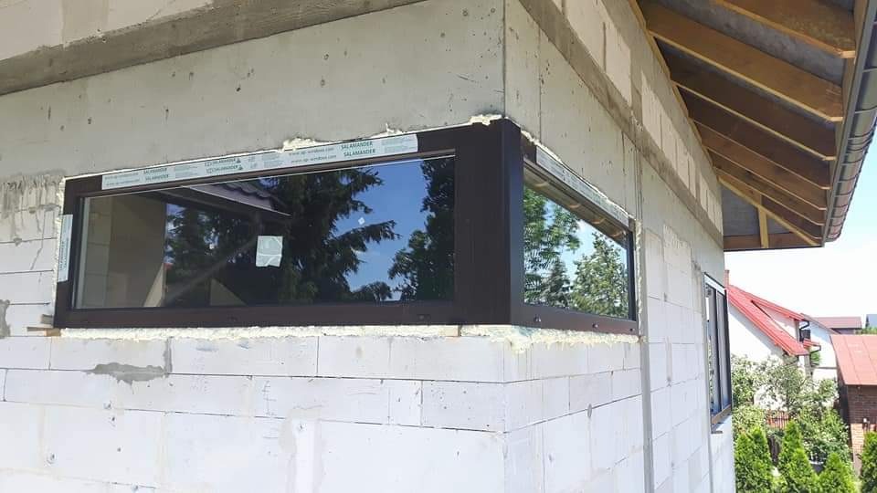 Montaż brązowych okien w surowym murze z betonu komórkowego, widoczne taśmy zabezpieczające ramę.