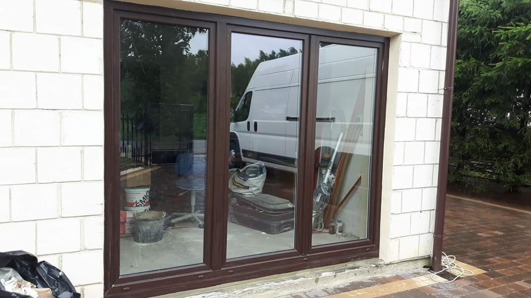 Brązowe trzyskrzydłowe okno z widocznym w odbiciu białym busem, narzędziami i materiałami budowlanymi, zamontowane w surowej ścianie z białego bloczka.