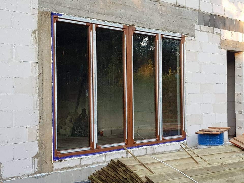 Montaż czteroskrzydłowego okna w surowym murze z betonu komórkowego, zabezpieczonego niebieską taśmą malarską.