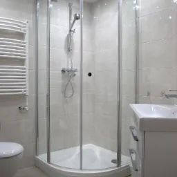 Jasna łazienka po remoncie: kabina prysznicowa narożna z chromowanymi profilami, biała umywalka z szafką i grzejnik drabinkowy na tle beżowych płytek.