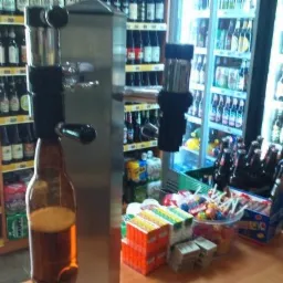 Nalewak do piwa z butelką, w tle półki z piwami i lodówka z napojami. Dodatkowo słodycze na ladzie. Widok z perspektywy klienta w sklepie.