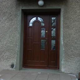 Brązowe drzwi wejściowe z szybkami i łukowym przeszkleniem, zamontowane w szarej, chropowatej ścianie budynku, widoczna rynna i lampa nad drzwiami.