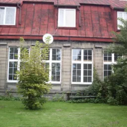 Fasada budynku z czerwonym dachem i białymi oknami, widoczne drzewa i trawnik, na dachu antena satelitarna.