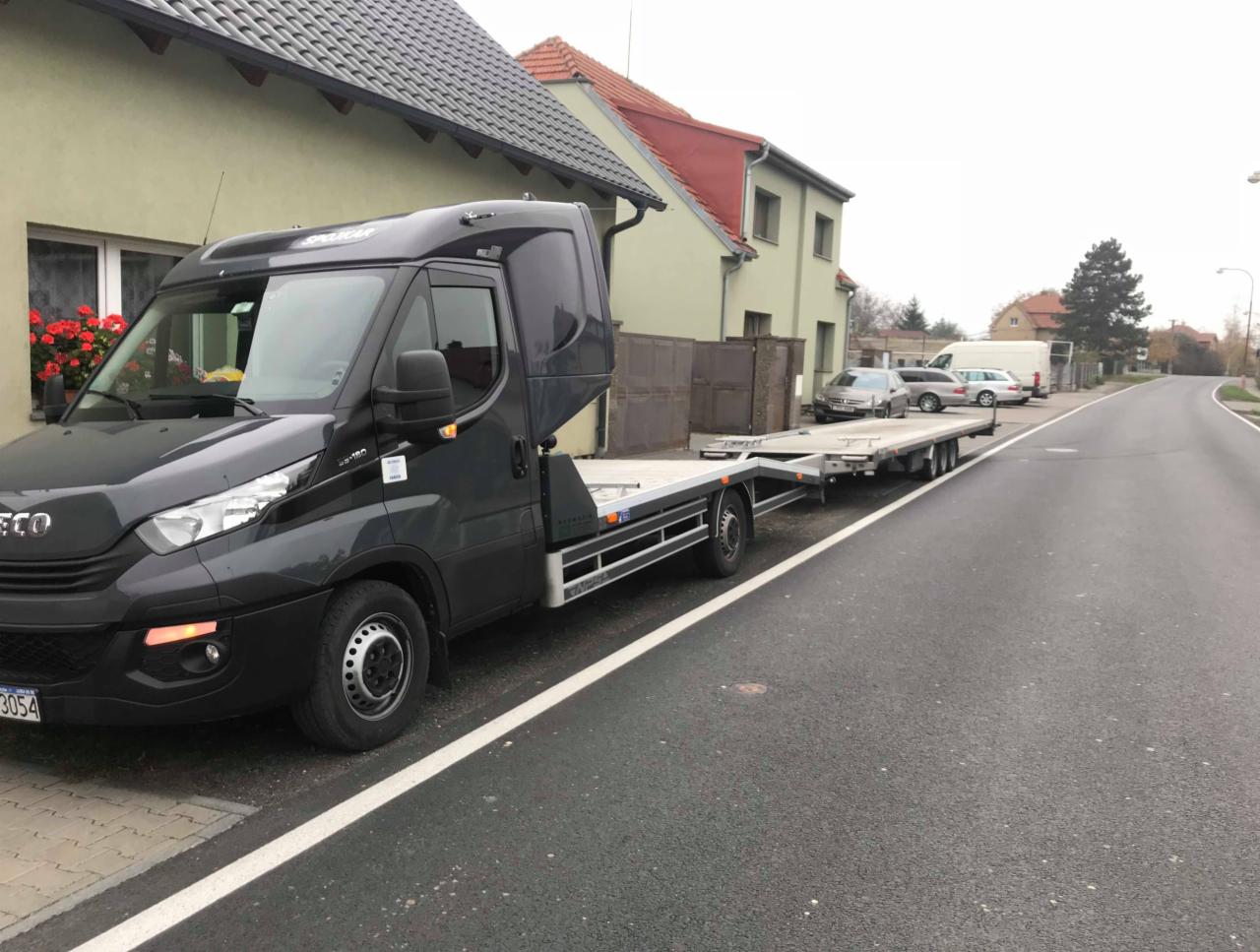 Ciemnoszara laweta Iveco z pustą platformą na ulicy w zabudowie miejskiej, widoczne zaparkowane samochody w tle.