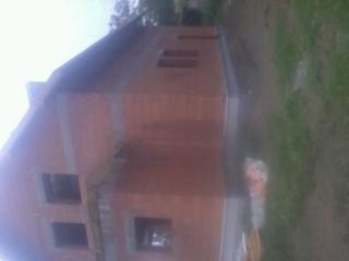 Surowy, niewykończony dom z czerwonej cegły, widok z boku, z oknami bez ram, na tle zieleni. Budynek w trakcie budowy, z widocznymi elementami konstrukcyjnymi.