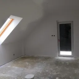 Pokój na poddaszu w trakcie remontu, widoczne okno dachowe, drzwi z roletą wewnętrzną i ślady po szpachlowaniu na podłodze, białe ściany przygotowane pod malowanie.
