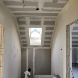 Montaż konstrukcji sufitu podwieszanego z oknem dachowym: widoczne profile i płyty w trakcie instalacji, pracownik sprząta podłogę w pomieszczeniu z surowymi ścianami.