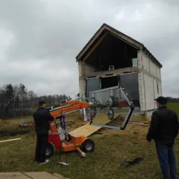 Montaż dużego okna przez dw&oacute;ch mężczyzn przy użyciu pomarańczowego podnośnika na budowie domu w stanie surowym zamkniętym.