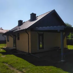 Dom jednorodzinny w trakcie budowy, z widocznymi czarnymi oknami i drzwiami zewnętrznymi, elewacja w stanie surowym, dach pokryty blachą, widoczne rynny, trawnik przed budynkiem.