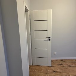PROCOMPLEX INTERIOR SP.Z O.O. - Białe, częściowo otwarte drzwi z czarną klamką i poziomymi frezami, na tle szarej ściany i drewnopodobnej podłogi, widoczne gniazdko elektryczne.