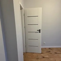 Białe, częściowo otwarte drzwi z czarną klamką i poziomymi frezami, na tle szarej ściany i drewnopodobnej podłogi, widoczne gniazdko elektryczne.