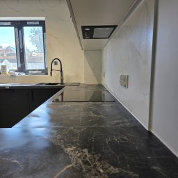 PROCOMPLEX INTERIOR SP.Z O.O. - Nowoczesna kuchnia z czarnym blatem imitującym marmur, białymi płytkami na ścianie i czarną armaturą. Widoczne okno z widokiem na zewnątrz i fragment zabudowy kuchennej.