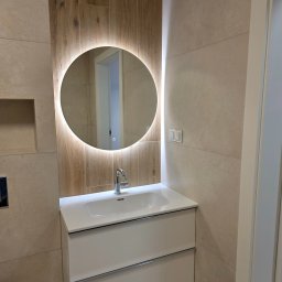 PROCOMPLEX INTERIOR SP.Z O.O. - Nowoczesna łazienka po remoncie: beżowe płytki, okrągłe lustro z podświetleniem LED, biała szafka z umywalką i chromowana bateria. Widoczna wnęka w ścianie.