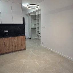 PROCOMPLEX INTERIOR SP.Z O.O. - Nowoczesne wnętrze: beżowe płytki podłogowe, drewniana zabudowa kuchenna z czarnym blatem i białe szafy wnękowe. Okrągła lampa sufitowa dodaje elegancji.