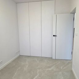 PROCOMPLEX INTERIOR SP.Z O.O. - Minimalistyczne wnętrze: gładkie, białe ściany, szafa w zabudowie, drzwi z czarnym akcentem i podłoga z płytek imitujących marmur. Nowoczesny design.