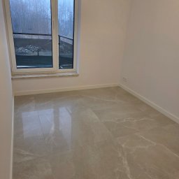 PROCOMPLEX INTERIOR SP.Z O.O. - Jasne, marmurowe płytki podłogowe w pustym pokoju z oknem balkonowym. Widok na zewnątrz ukazuje drzewa i gruzowisko. Minimalistyczne wykończenie wnętrza.