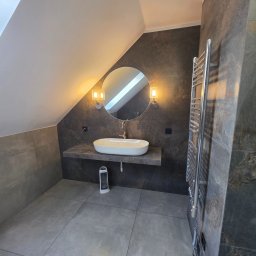 Nowoczesna łazienka na poddaszu w Rydułtowach. Umywalka nablatowa, okrągłe lustro z oświetleniem, grzejnik drabinkowy i szare płytki tworzą spójną całość.