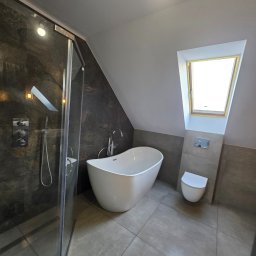 Nowoczesna łazienka na poddaszu z kabiną prysznicową, wolnostojącą wanną i toaletą. Ciemne płytki kontrastują z bielą armatury i jasnym oknem dachowym.