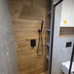 PROCOMPLEX INTERIOR SP.Z O.O. - Nowoczesna kabina prysznicowa z drewnopodobnymi płytkami i czarną armaturą. Minimalistyczny design z szarymi półkami i elementami wykończenia.