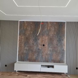 PROCOMPLEX INTERIOR SP.Z O.O. - Nowoczesny salon z dekoracyjną ścianą z paneli pionowych i abstrakcyjną tapetą. Biała szafka RTV i podświetlany sufit tworzą spójną całość.