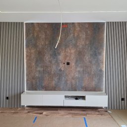 PROCOMPLEX INTERIOR SP.Z O.O. - Wykończona ściana z dekoracyjnymi lamelami po bokach i panelem imitującym rdzę w centrum. Biała szafka RTV podwieszona. Widoczne kable i zabezpieczona podłoga.