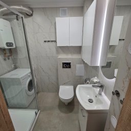PROCOMPLEX INTERIOR SP.Z O.O. - Kompletna, jasna łazienka z kabiną prysznicową, pralką, umywalką z szafką, toaletą i szafkami wiszącymi. Marmurowe płytki ścienne i szara podłoga. Oświetlenie LED.