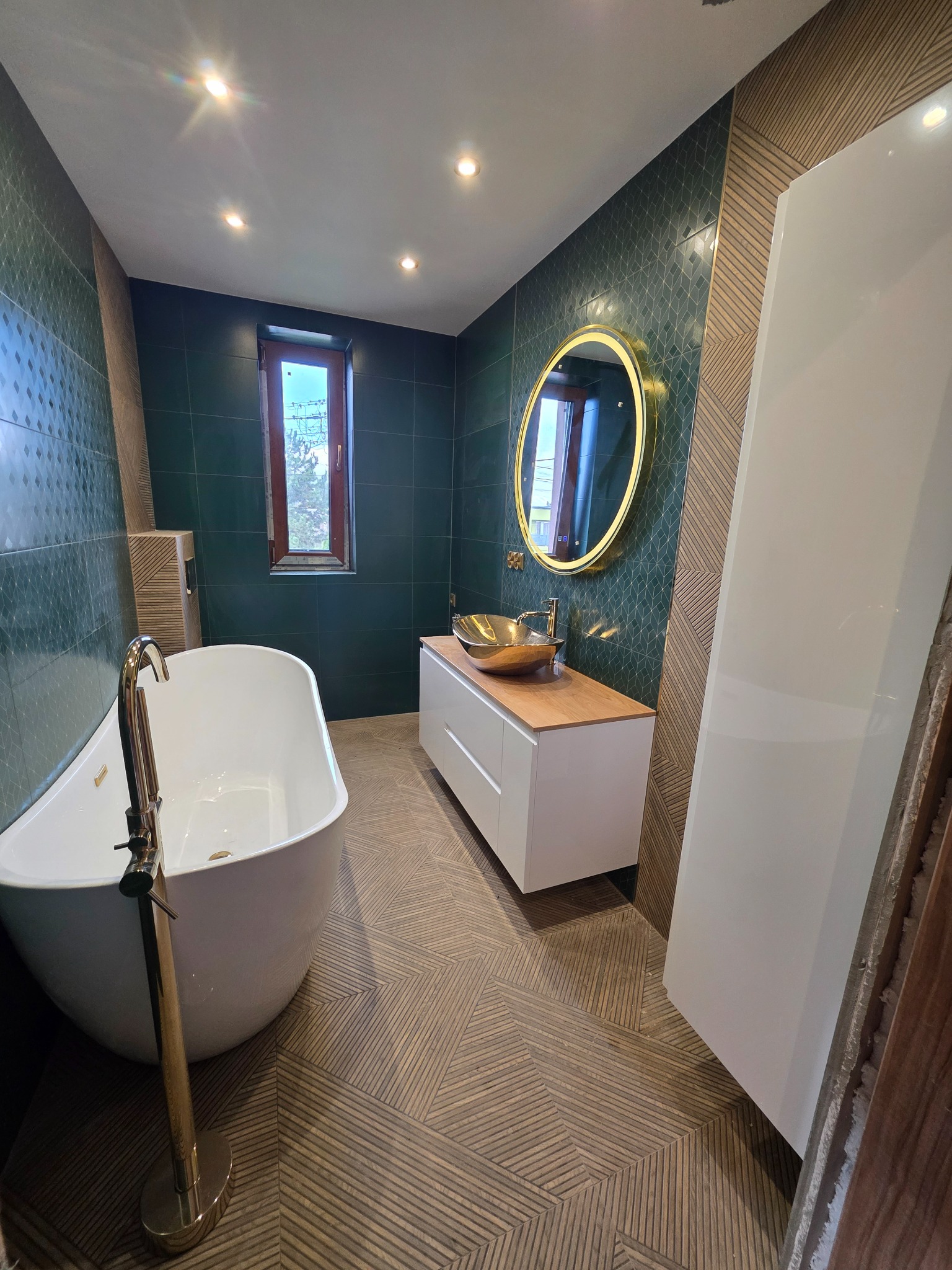 Nowoczesna łazienka z zielonymi płytkami, złotym lustrem i umywalką. Parkiet ułożony w jodełkę. Wolnostojąca wanna i biała szafka pod umywalką. Okno z drewnianą ramą.