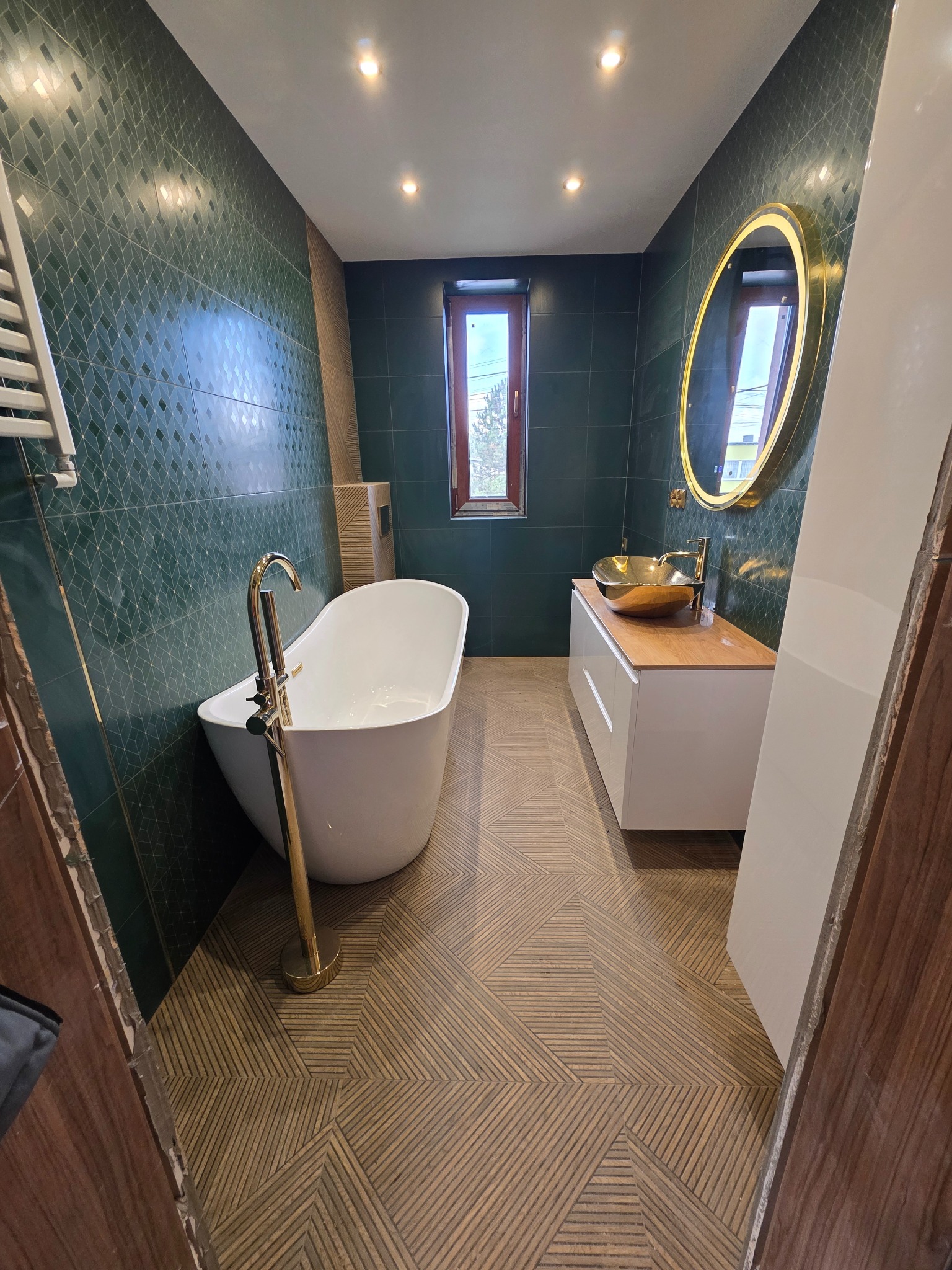 Nowoczesna łazienka z zielonymi płytkami, wolnostojącą wanną z złotą armaturą i umywalką na drewnianym blacie. Geometryczny wzór płytek podłogowych.