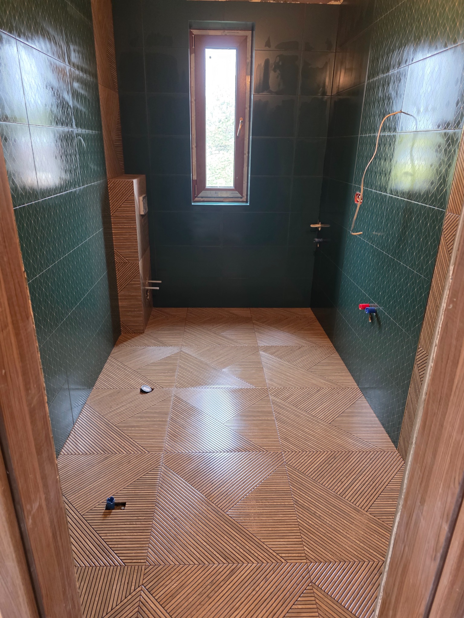 Wykończona łazienka z zielonymi, błyszczącymi kaflami na ścianach i wzorzystą drewnopodobną podłogą. Widok zza drewnianej futryny drzwi.