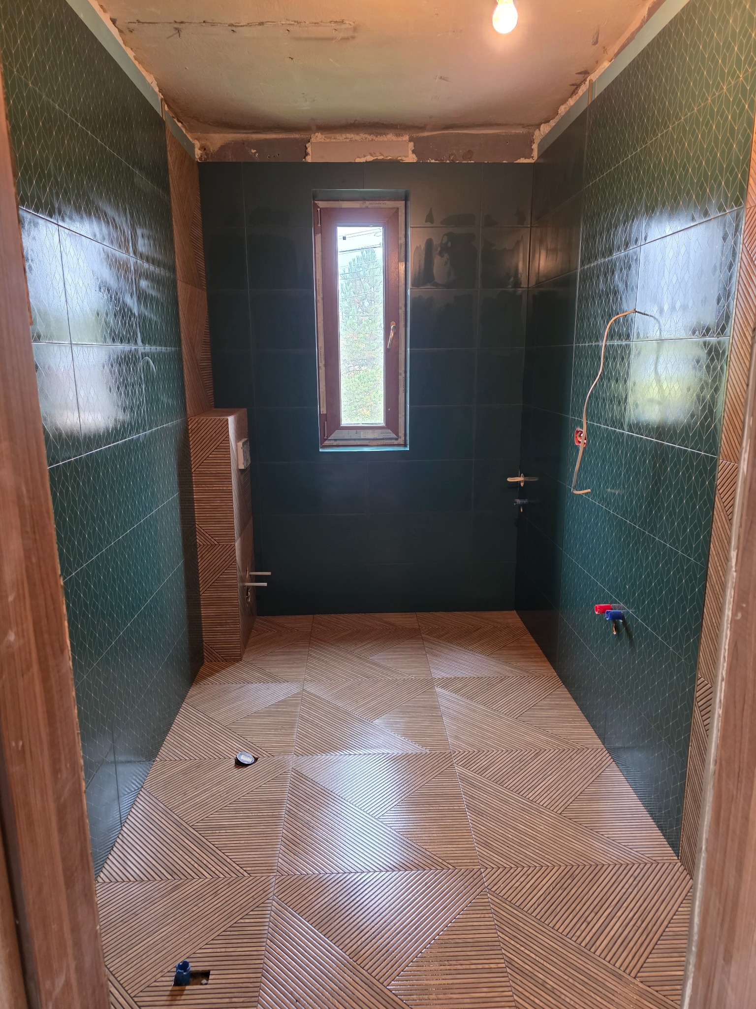 Łazienka w trakcie remontu: ułożone płytki podłogowe z wzorem jodełki, ciemnozielone kafelki na ścianach, widoczne instalacje, okno w tle, surowy sufit.