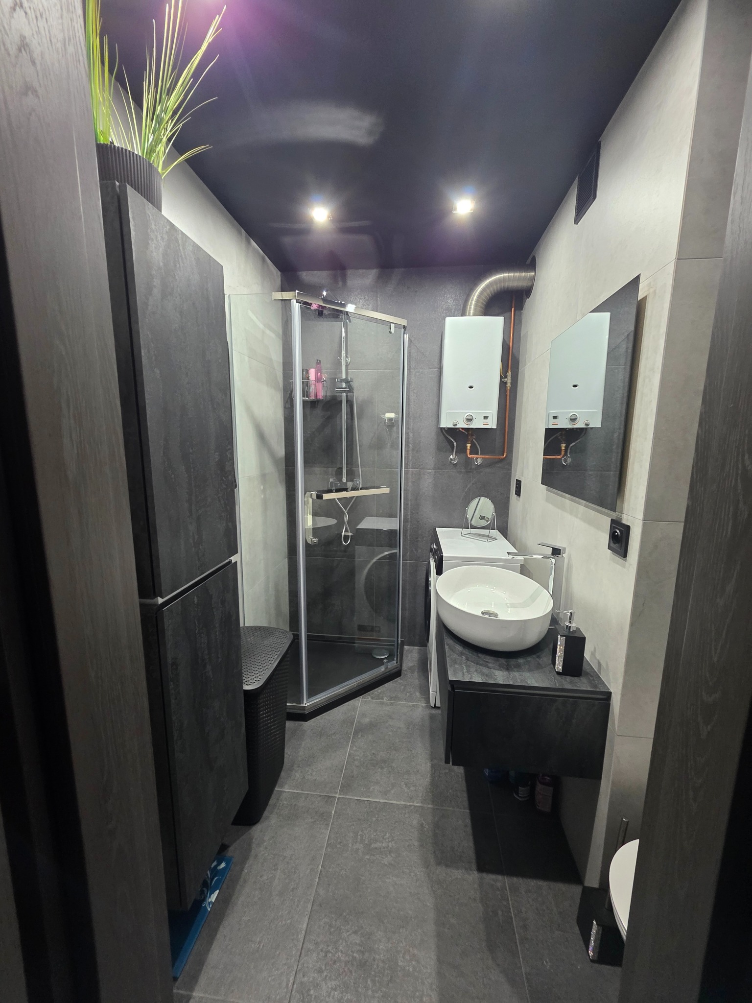 Nowoczesna, szaro-czarna łazienka po remoncie: kabina prysznicowa, umywalka nablatowa, szafka, pralka i bojler. Minimalistyczny design z rośliną doniczkową.