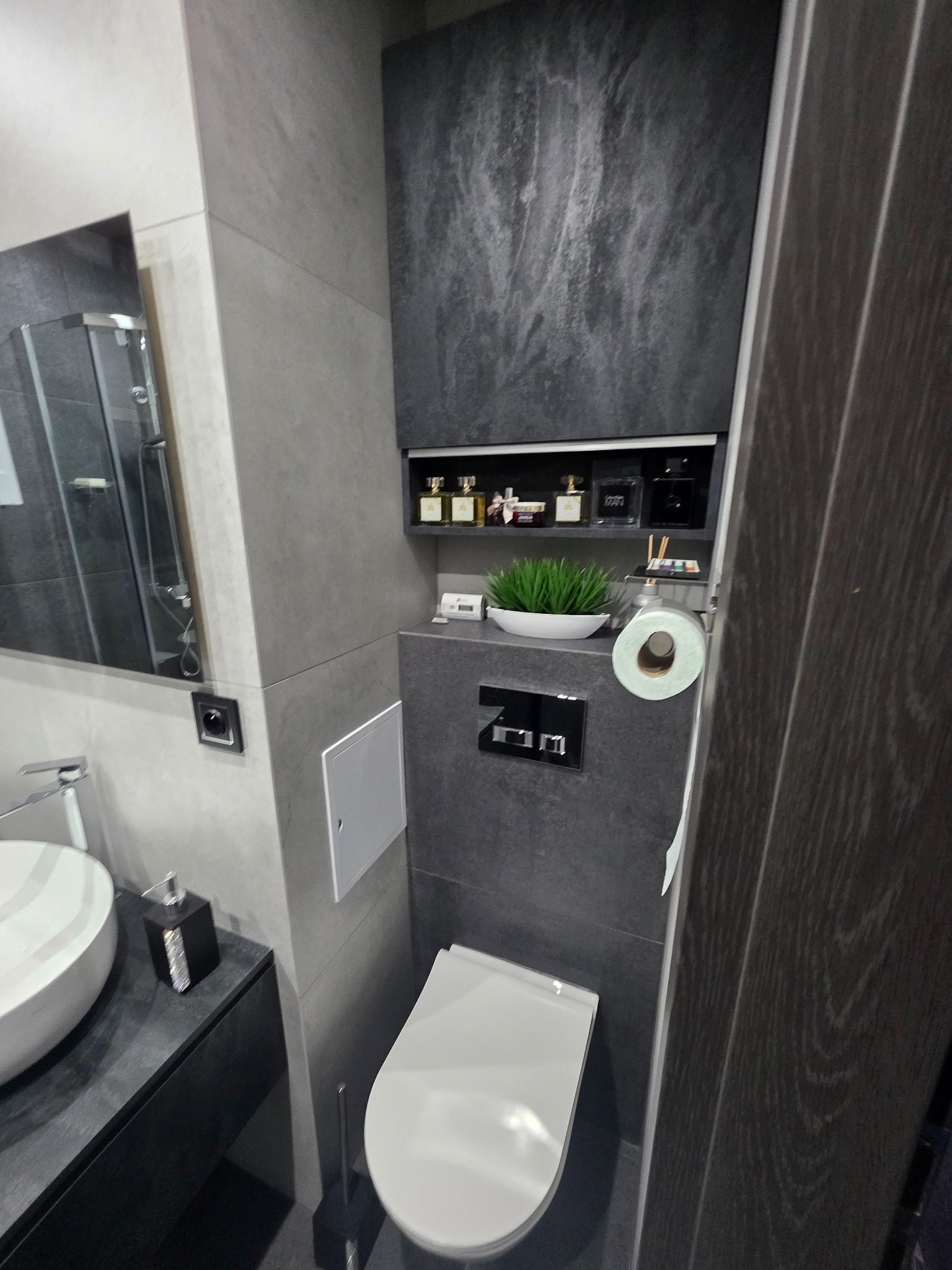 Nowoczesna łazienka w szarościach z toaletą w zabudowie, umywalką na blacie i lustrem. Na ścianie półka z dekoracjami. Minimalistyczny design.