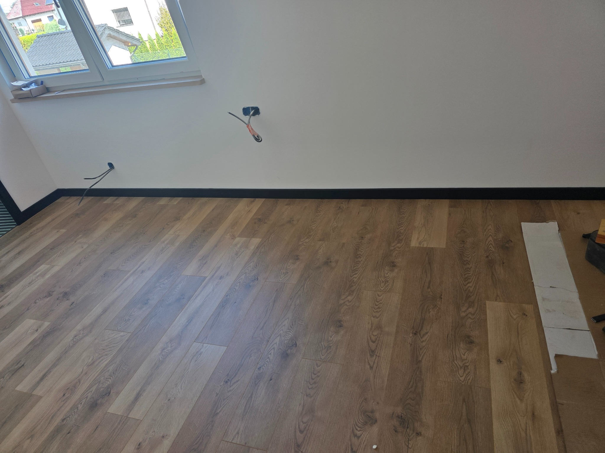 Ułożone panele podłogowe w pokoju na poddaszu, widoczne kable elektryczne wystające ze ściany, listwy przypodłogowe. Częściowo widoczne narzędzia.