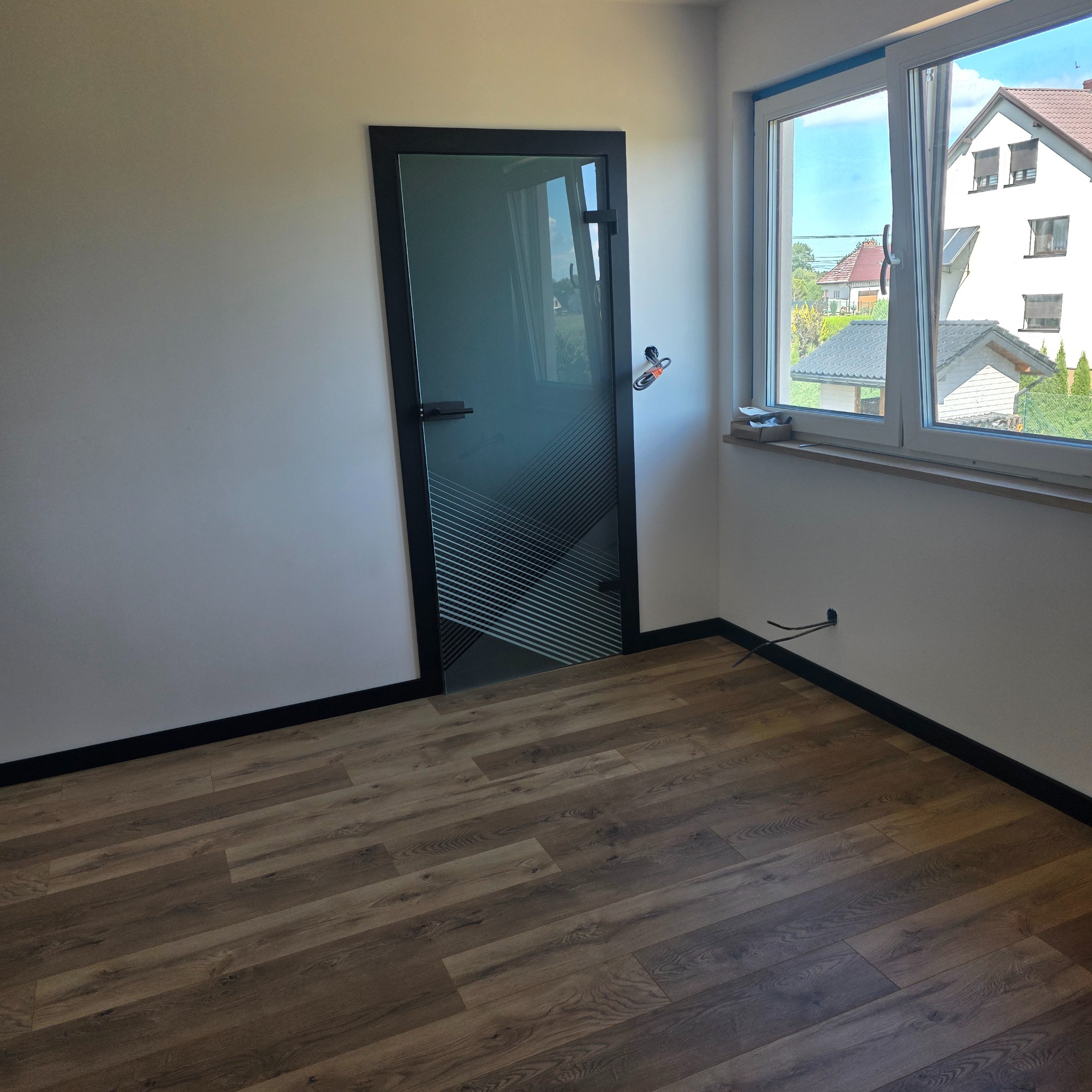 Pokój po remoncie: brązowe panele podłogowe, białe ściany z czarnymi listwami, nowoczesne drzwi z geometrycznym wzorem i okno z widokiem na dom.