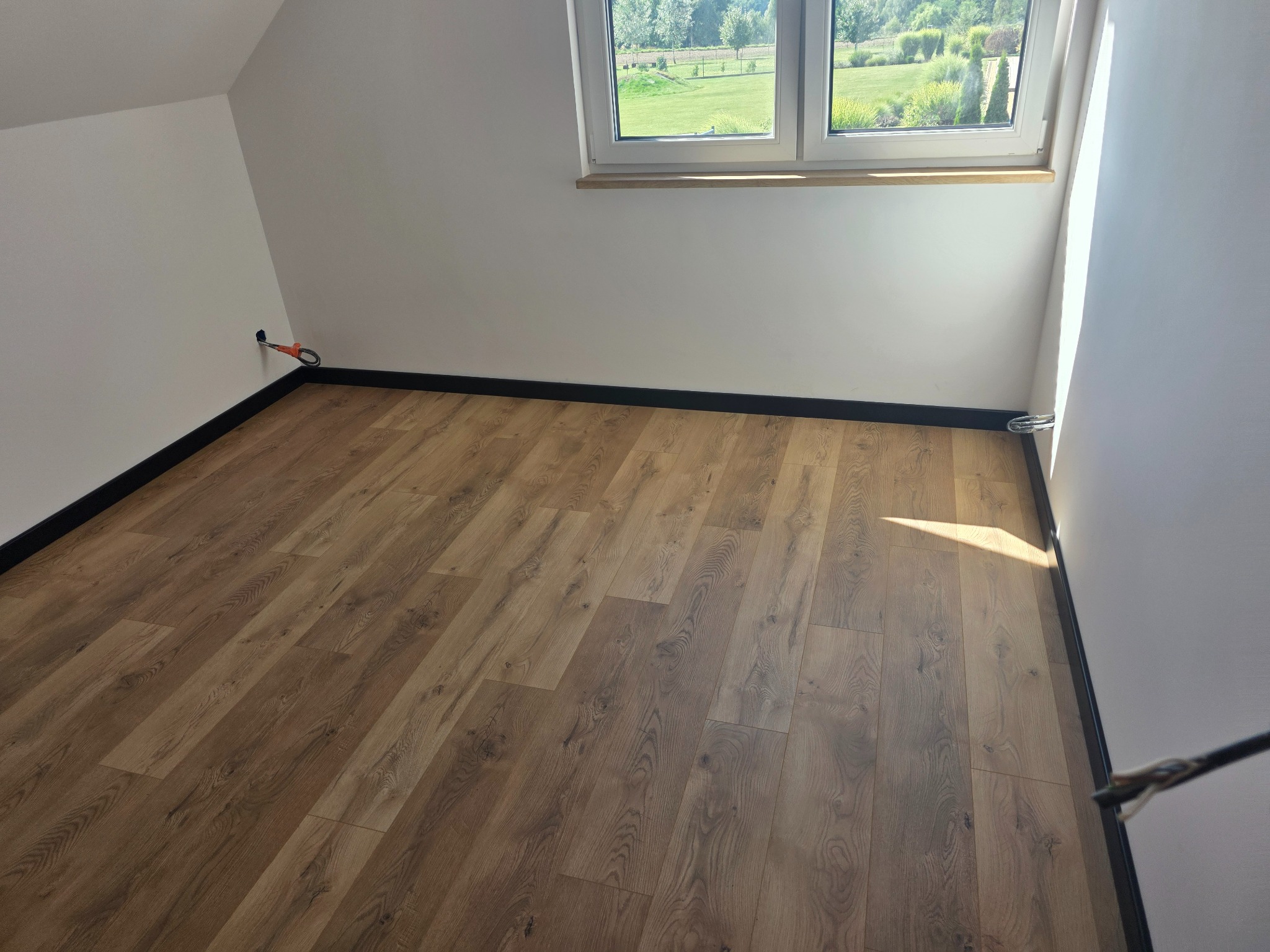 Jasne panele podłogowe ułożone w pokoju na poddaszu z oknem i czarnymi listwami przypodłogowymi. Widoczne gniazdko elektryczne i fragment narzędzia.