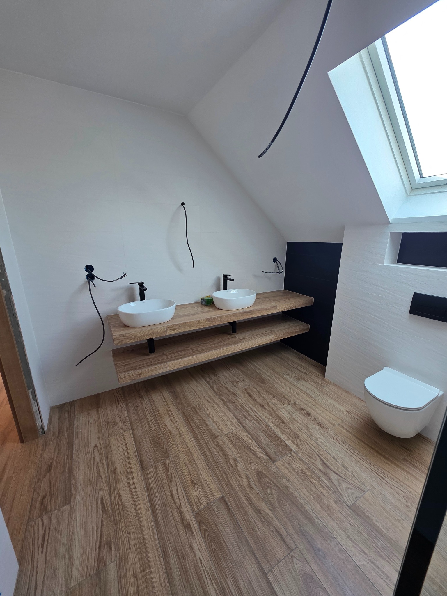 Nowoczesna łazienka na poddaszu z drewnianą podłogą i blatem pod umywalki. Uwagę przykuwa okno dachowe wpuszczające naturalne światło i minimalistyczna toaleta.
