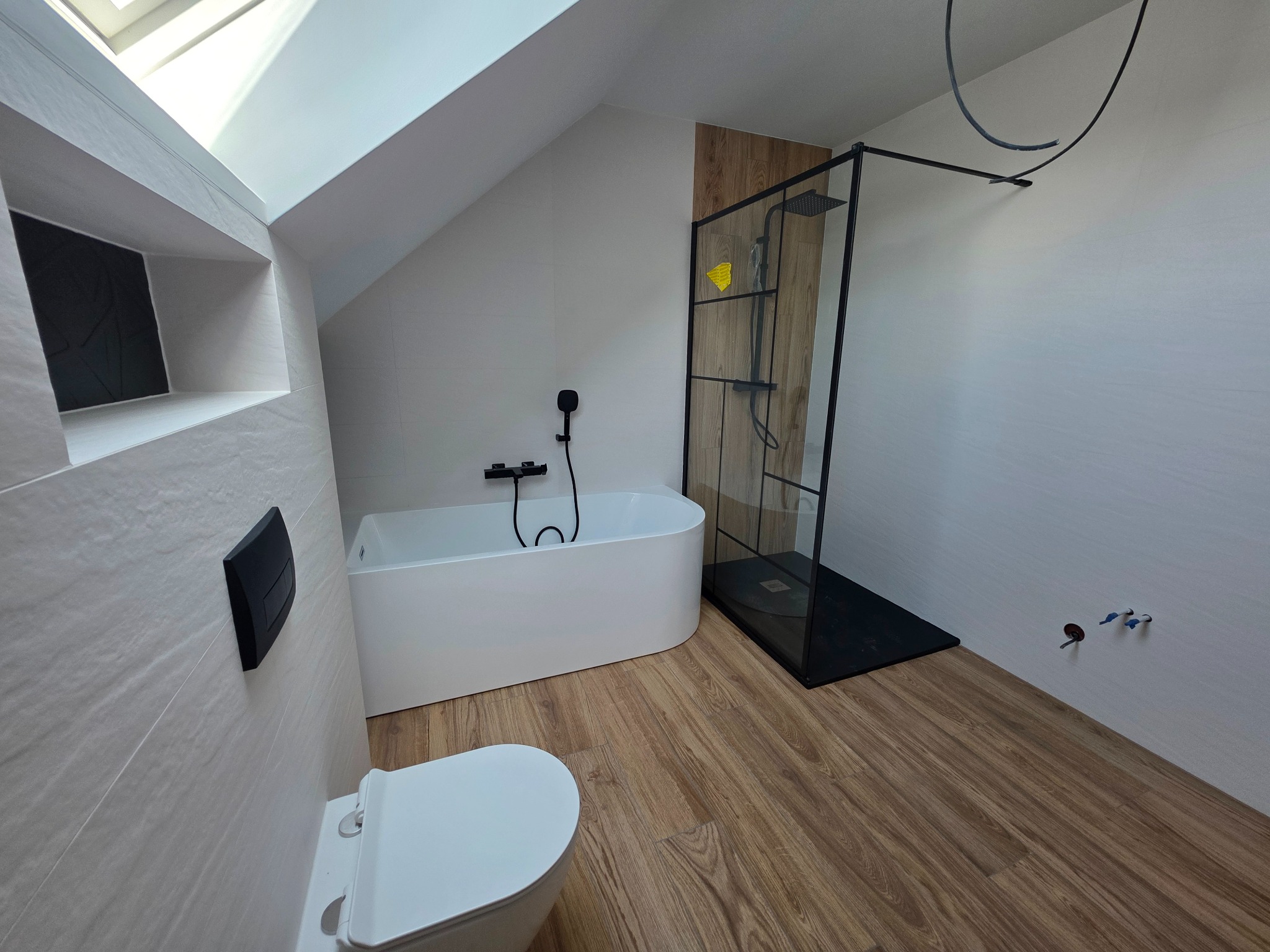 Nowoczesna łazienka na poddaszu: biała wanna, kabina prysznicowa z czarną ramą, drewnopodobne panele na podłodze i ścianie, okno dachowe. Minimalistyczny design.