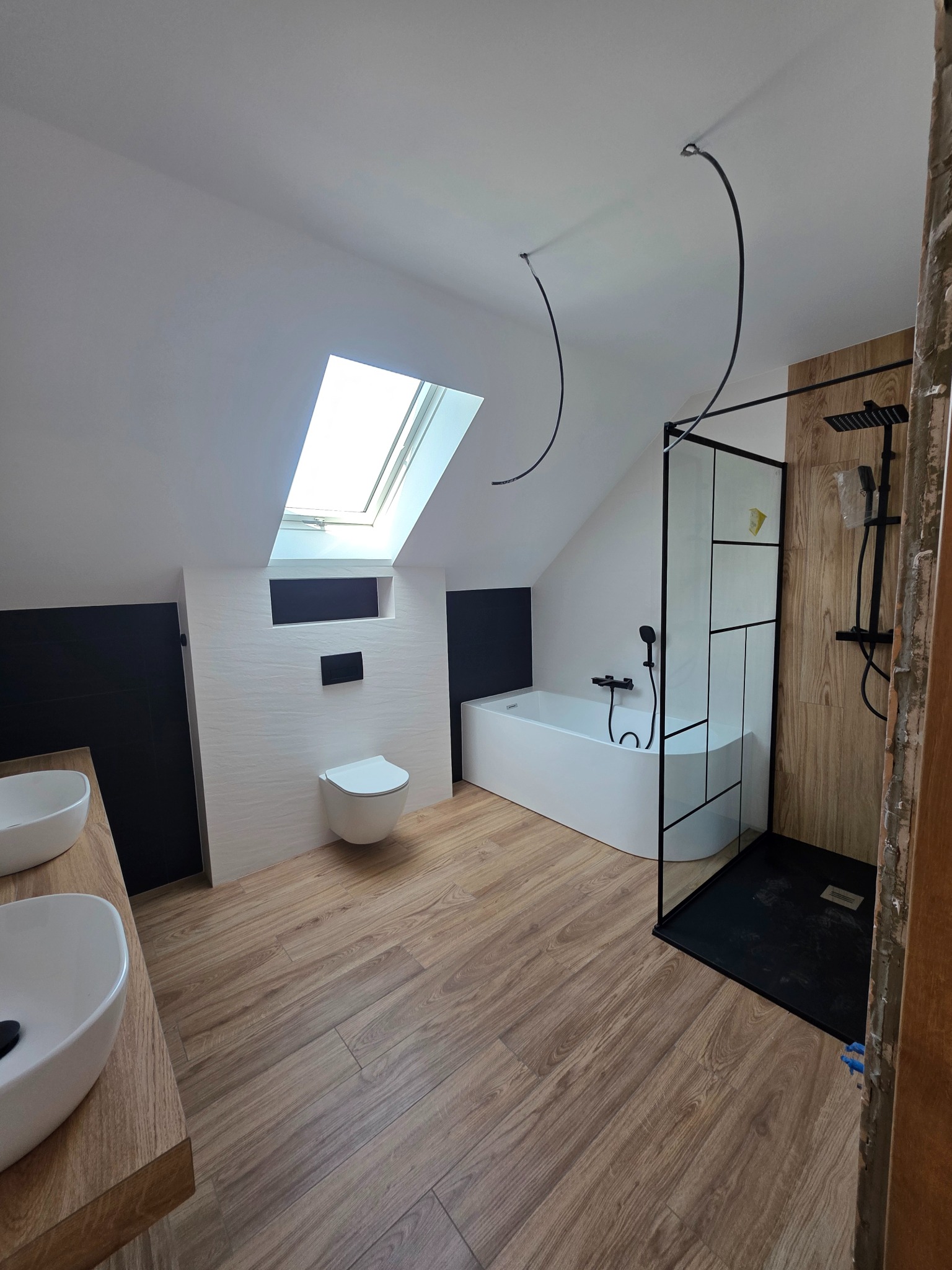Nowoczesna łazienka na poddaszu ze skośnym oknem dachowym. Uwagę przykuwa czarna armatura i kabina prysznicowa z czarnymi profilami. Podłoga wyłożona drewnopodobnymi płytkami.