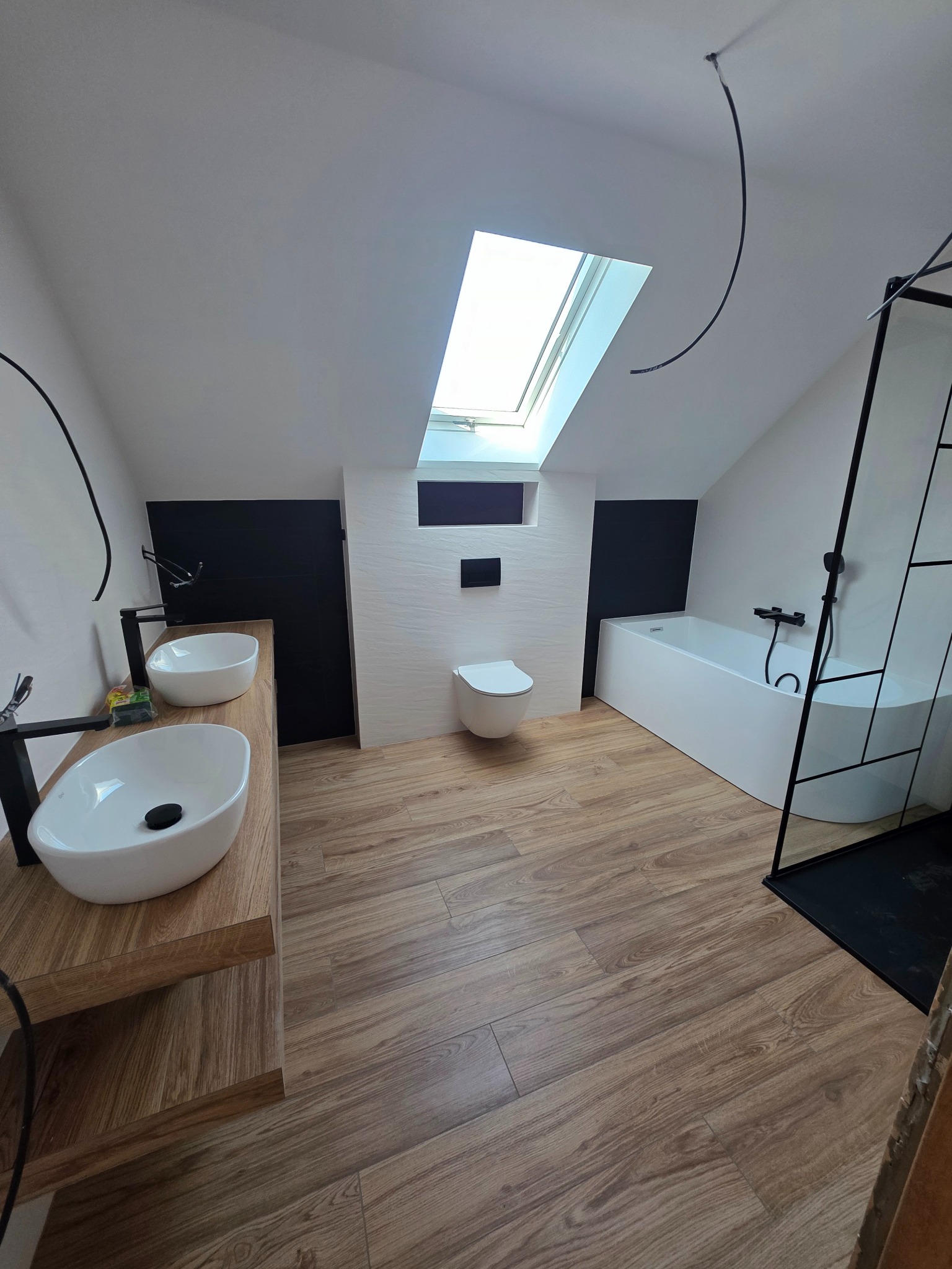 Nowoczesna łazienka na poddaszu z drewnopodobną podłogą, białą armaturą i czarnymi akcentami, oświetlona światłem dziennym z okna dachowego.